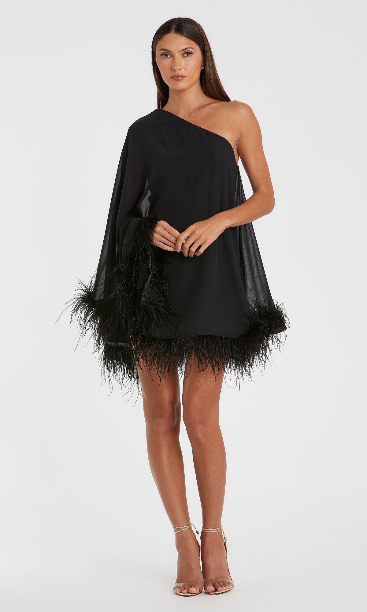 A woman models the Mac Duggal 11623 short wedding guest dress, featuring a black one-shoulder silhouette with a feather-trimmed hem, styled with nude heels—an elegant choice against a white backdrop.